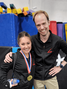 Coach Adam with his arm around a female athlete. Both are smiling and wearing black jackets with a red Legacy "L" logo on them. The female athlete has two gold medals around her neck, and is holding a bottle of water. Behind the two, is a stack of gymnastic mats and blocks in various colors.